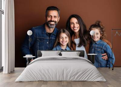 Happy family of four, including an adult man, an adult woman, and two young girls, posing against a plain brown background. They are all smiling and wearing casual clothing. Wall mural