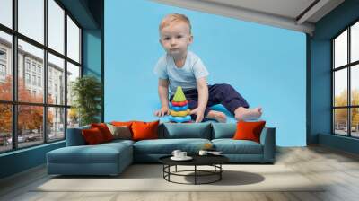 Little boy collects a toy pyramid sitting on a blue background, copy space. Wall mural