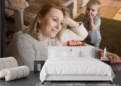 Adult blond woman sit on sofa at home relaxing while small daughter play alone Wall mural