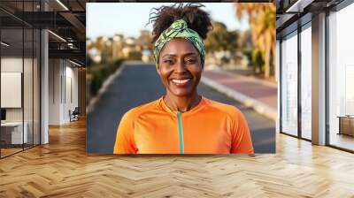 Smiling woman with afro hair wearing an orange athletic shirt and a green headband, standing outdoors on a pathway in a park setting. Wall mural