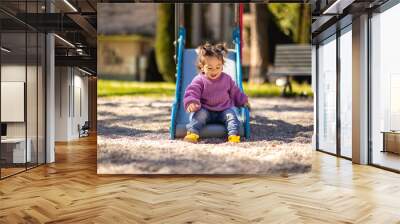 Sweet caucasian little toddler girl with two tails having fun smiling widely sliding down the yellow hill head first in the playground with violet sweeter sleeveless jacket jeans Wall mural