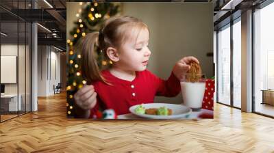 the child eats cookie and drinks milk on the background of a Christmas tree with lights. children's xmas dinner Wall mural