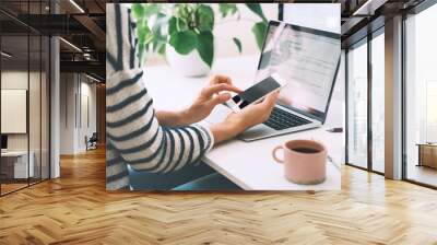 Woman using smartphone while working on laptop in home office. Wall mural