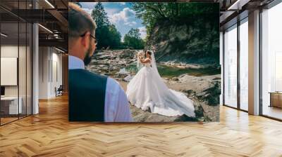 Romantic outdoor wedding shot of the fashionable newlywed couple hugging at the river bank. Bride and groom kiss and hug each other behind beautiful landscape. After wedding ceremony. Wall mural