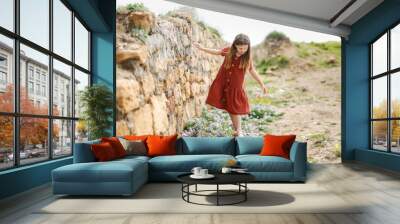 Cute caucasian child girl in a summer dress walks barefoot over stones and wild flowers. Summer holidays, childhood and natural wild plants and flowers among the stones Wall mural