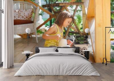 cute caucasian girl child in a yellow short-haired t-shirt watering flowers in pots on the veranda, caring for plants and helping children with household chores Wall mural