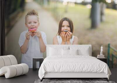 Cute romantic couple of kids with donuts. Brother and sister twins eat pink and chocolate donuts in summer outdoor Wall mural