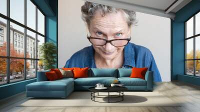 Close up of senior woman with blue eyes peering over glasses against neutral background (selective focus) Wall mural