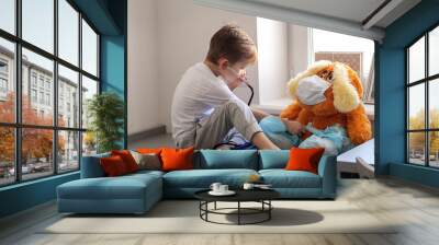 child plays a doctor. The boy listens to the breath of a toy to a large dog with a stethoscope. A toy in a medical bandage and shoe covers on legs. Wall mural