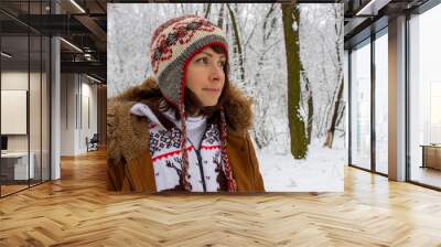 Thoughtful girl in knitted cozy wear in snowy winter forest. Winter weather and clothes concept. Happy woman in deer sweater and nepalese hat in frozen park. Christmas background. Positive lifestyle. Wall mural