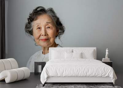 A asian woman with white hair and a white shirt is smiling. She is looking at the camera. The image has a calm and peaceful mood. Asian elderly woman in white blouse isolated on light grey background Wall mural