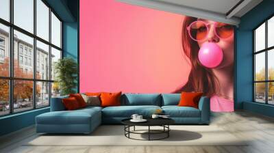 A beautiful young funny girl in pink clothes on a pink background inflates a large pink bubble gum ball in her mouth. Girl in pink glasses. Place for text. Wall mural