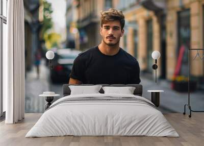 A man in a black shirt is standing on a sidewalk in front of a building. He has a beard and is looking at the camera. Male model in a classic black cotton T-shirt on a city street Wall mural