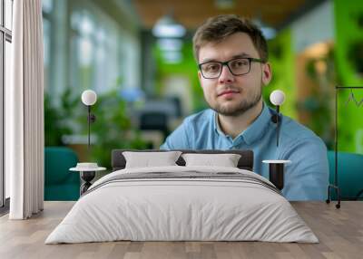 A man wearing glasses and a blue shirt sits in a green chair. He is looking directly at the camera. brutal handsome young man of European type, average man with broad shoulders Wall mural