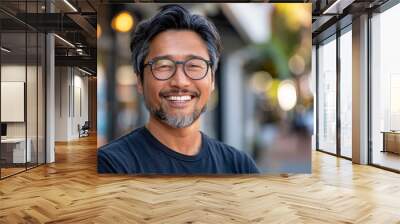 A man with glasses is smiling and wearing a blue shirt. He is standing in front of a building with a backpack on his back. Happy satisfied asian man wearing glasses portrait outside. Wall mural