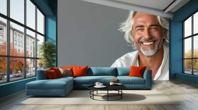 A man with white hair and a white shirt is smiling. He is sitting on a grey surface. Attractive 50 year old smiling man with medium white hair in cream-colored clothes against a gray background. Wall mural