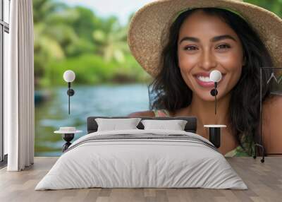 A woman wearing a straw hat and a floral dress is smiling at the camera. She is sitting in a boat on a river Wall mural