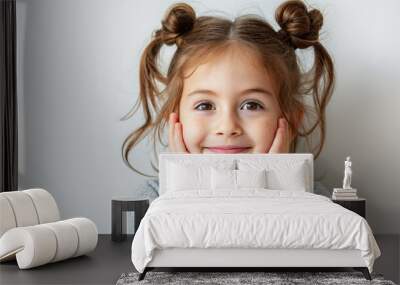 A young baby girl with brown hair and a smile on her face. She is wearing a gray shirt and has her hands on her face. Cute young child with prominent protruding ears on a light background. Wall mural