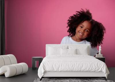 A young girl with curly hair is smiling at the camera. The pink background adds a warm and inviting atmosphere to the image Wall mural