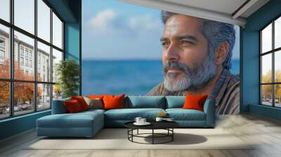 Man with a beard and gray hair is sitting on beach looking out at the ocean. handsome 50 year old Indian man, he is sitting by the shore, he is wearing a flannel shirt, he is holding a fishing rod, Wall mural