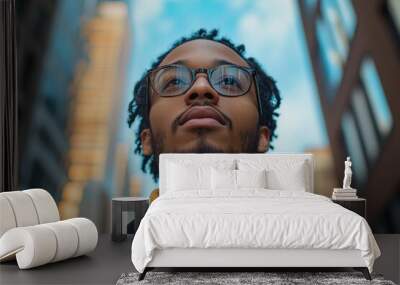 Man with glasses and a beard is looking up at the sky. The image has a mood of contemplation and introspection. thoughtful man stands against a backdrop of modern high-rise buildings Wall mural