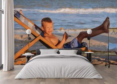 tanned baby boy resting and sunbathing in a deck chair on the sand by the blue sea Wall mural