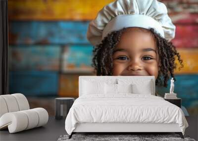 Young girl with curly hair is wearing a chef's hat and apron. She is smiling and looking at the camera. a cute child wearing a chef's hat smiling with a bright colored background Wall mural