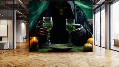 One witch in black clothes with a hood in a mask and black leather gloves holds two glasses with a green poisonous drink while sitting at a table with candles and pumpkins in a dark room, close-up sid Wall mural