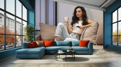 Pretty young smiling woman sitting on sofa at home watching TV with cup of hot drink. Attractive female resting on couch holding remote control in hand watching television Wall mural