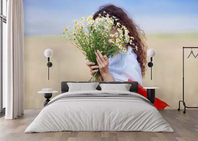 Woman with flag of Poland on shoulders holding bouquet of daisies in front of face in wheat field. Polish Flag Day. Independence Day. Travel and learn polish language concept. Selective focus. Wall mural