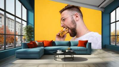 Creative closeup portrait of a young handsome bearded guy with glasses in a white t-shirt tastes a hamburger from a fast food on the yellow background. Wall mural