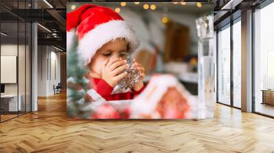 Happy funny toddler boy in a red Santa hat drinking filtered water from a glass in the kitchen. Holidays, health concept. Wall mural