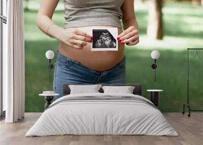 Happy young adult pregnant girl holds in her hands a snapshot of an ultrasonic study of a toddler walking in a park. Second trimester. Wall mural