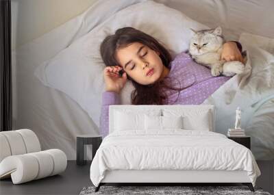 A little brunette girl in purple pajamas sleeps hugging a white charming cat lying on a white pillow on the bed. Wall mural