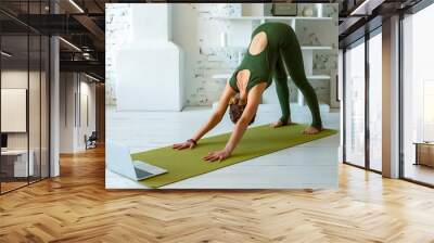 A slender woman in a green sports jumpsuit performs yoga exercise at home, in a light interior, Wall mural