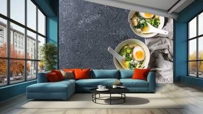 Two bowls with asian style soup with scrambled eggs, half of marinated egg, spring onion, spinach served with white spoons and textile over black texture concrete background. Top view with space Wall mural