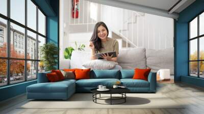 Beautiful attractive young Asian woman sitting on gray sofa and using digital tablet at living room in the morning. Wall mural