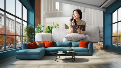 Beautiful attractive young Asian woman sitting on gray sofa and using digital tablet at living room in the morning. Wall mural