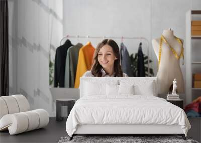 Portrait of tailor woman working on clothes in tailoring atelier. Attractive beautiful young female fashion designer work on laptop in workshop Wall mural