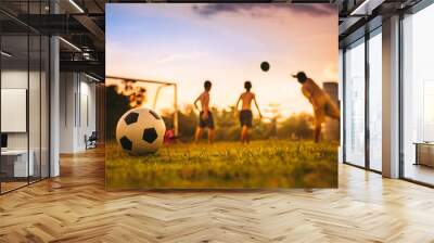 Silhouette action sport outdoors of a group of kids having fun playing soccer football for exercise at the green grass field in community rural area under the twilight sunset sky. Wall mural