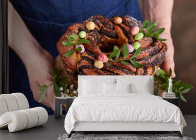 Man in a blue apron holding a freshly baked Easter cake with poppy seed filling and candies in a shape of eggs. Selective focus. Round yeast dough sweet bun. Wall mural