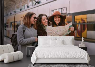 Three women travelers friends are at the train station checking the map of the city. They are laughing together while looking for a place to go. Travel concept Wall mural