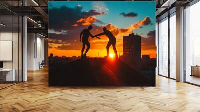 Urbex duo in silhouette, one pulling the other up on a rooftop edge, sunset city skyline, parkour challenge in urban setting, teamwork in focus Wall mural