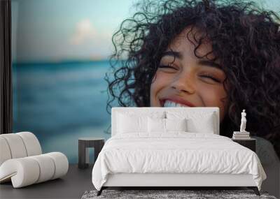 Cheerful portrait of young woman with curly hair, smiling joyfully by beach, capturing moment of happiness and warmth. ocean waves create serene backdrop Wall mural