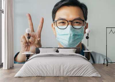 Handsome Asian doctor wearing white coat and blue shirt, smiling and showing peace sign. His friendly demeanor conveys warmth and professionalism Wall mural