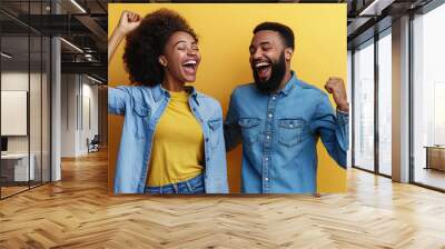 Joyful couple in matching denim outfits celebrating together against vibrant yellow background, expressing happiness and excitement Wall mural