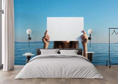 woman holds blank white sign in front of her face against serene ocean backdrop, evoking sense of mystery and calm. clear blue sky and tranquil water enhance peaceful atmosphere Wall mural