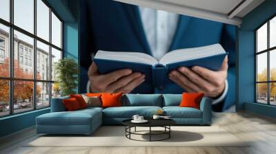 A person in a suit holds an open book, focusing on the pages while standing against a neutral background. Wall mural