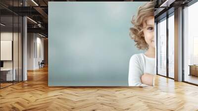 Confident child with curly hair, wearing a light sweater, smiling gently, arms crossed, set against a soft blurred background creating a warm and inviting atmosphere Wall mural