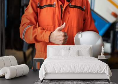 A repairman in orange coverall is thumbing up and holding white safety helmet with blurred background of garage workshop. Safe working practice in industrial concept. Wall mural